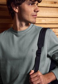 Muž v světle zelené mikině drží černý popruh od batohu, stojí před dřevěnou stěnou s latěmi a dívá se na stranu.