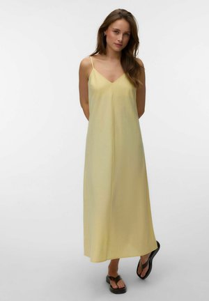 Kvinde med langt brunt hår iført en lys gul ærmeløs maxikjole og sorte platformssandaler, stående mod en ensfarvet lys baggrund.