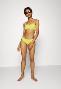 Conjunto de bikini amarelo com tecido texturizado; inclui um top tipo bralette com alças ajustáveis e uma parte inferior de cintura baixa combinando. A modelo usa óculos de sol.