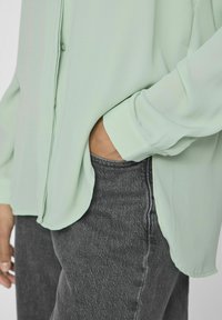 Camicia button-up verde chiaro con una texture liscia e maniche lunghe, abbinata a jeans grigi scuri, che mostrano una vestibilità casual.