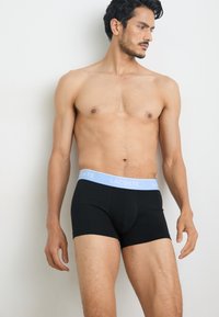 Black boxer shorts with a light blue elastic waistband featuring the Lacoste logo. The fabric appears smooth with a close-fitting design.
