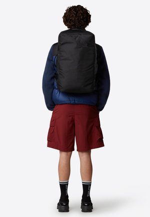 Sort rygsæk med lynlås, rektangulær form, polstret ryg og justerbare stropper, båret over en blå polstret jakke og røde shorts.