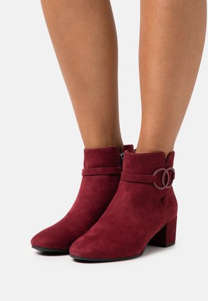Botas de tobillo de ante burdeos con punta redondeada, cuello con borde festoneado, cierre lateral y un adorno de doble círculo decorativo en la correa. Tacón cuadrado.
