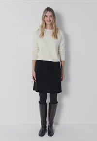 Pull en tricot blanc, jupe en suède noire, collants noirs, bottes en cuir noir montantes. Debout contre un fond neutre.