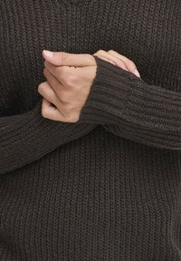 Donkerbruin gebreid sweater met een gestructureerd patroon. Close-up toont ribgebreide manchetten en handen die op de mouwen zijn geklemd, waardoor het materiaal wordt benadrukt.