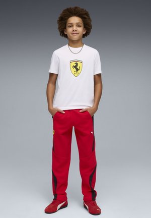 Maglietta bianca con logo Ferrari giallo, abbinata a pantaloni jogger rossi con dettagli laterali neri. Scarpe sportive rosse completano il look.