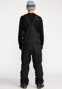 Zwarte bibtrok met verstelbare banden, zij- en achterzakken, en een relaxte pasvorm. Draagt over een zwart longsleeve shirt en een grijze gebreide muts.