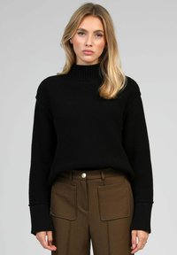 Femme portant un pull à col roulé côtelé noir et un pantalon marron avec de grandes poches avant, se tenant devant un fond gris uni.