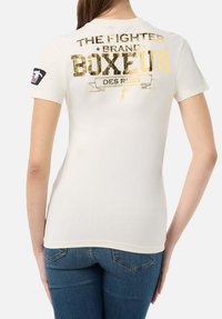 Weißes Baumwoll-T-Shirt mit goldfoliertem Text auf der Rückseite: "THE FIGHTER BRAND BOXEUR DES RUES" sowie einem dekorativen Banner und einem Logo-Patch am Ärmel.