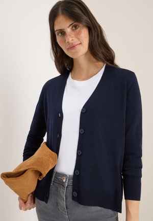 Navy cardigans met lange mouwen, knoopsluiting en geribde boorden. Gecombineerd met een wit shirt en met een tan suède clutch in de hand.