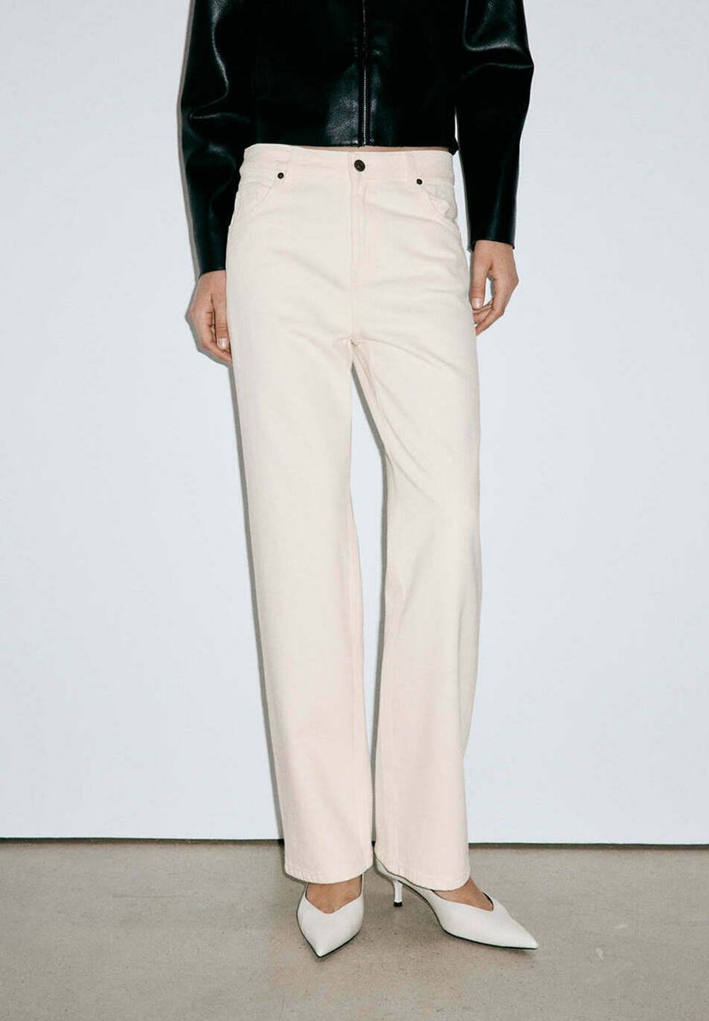 Femme portant un pantalon crème taille haute à jambes larges, un haut noir à manches longues, et des chaussures à talons blancs à bout pointu, debout devant un fond uni.