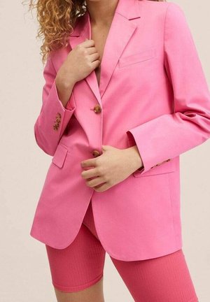 Person, die einen hellpinkfarbenen Blazer mit braunen Knöpfen und passende pinkfarbene gerippte Radlerhosen vor einem einfarbigen Hintergrund trägt.