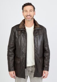 Man die glimlacht en een donkerbruine leren jas met een bontkraag draagt, een crèmekleurige trui en lichtgrijze jeans tegen een effen achtergrond.