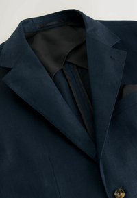 Blazer bleu marine avec col cranté, doublure intérieure noire et fermeture par un seul bouton, présenté plié sur une surface blanche.
