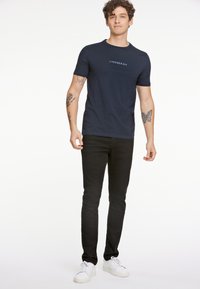 T-paita tummansinisestä puuvillasta, jossa on vaaleansinisellä painettu "LINDBERGH" -logo, yhdistettynä mustiin farkkuihin ja valkoisiin lenkkareihin. Malli seisoo.