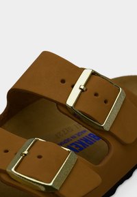 Καφέ δερμάτινο σανδάλι με δύο ρυθμιζόμενες χρυσές μεταλλικές αγκράφες και διαμορφωμένο πέλμα, με μερική σήμανση Birkenstock στο εσωτερικό.