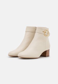 Beige kožené kotníkové boty s nízkým blokovým podpatkem, se širokým páskem a zlatou přezkou na kotníku. Hladký povrch a špičatá špička.