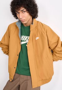 Nike bomber jacket in mustard yellow with gray lining; features elastic cuffs and hem, snap buttons, and a white logo on the chest.