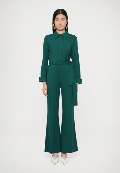 Tuta verde con scollo a collo, chiusura con bottoni sul davanti, design avvolgente, maniche lunghe e gambe ampie, abbinata a scarpe bianche a punta.