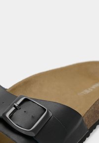 Gros plan d'une sangle de sandale en cuir noir avec boucle en métal sur une semelle intérieure couleur sable et une semelle en liège.