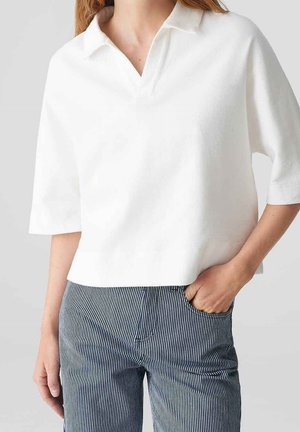 Persona que lleva una camisa blanca de manga corta con cuello y pantalones rayados verticales en azul y blanco, con la mano en el bolsillo, sobre un fondo liso.