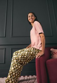 Šviesiai rožinė trumpomis rankovėmis marškinėliai; platėjančios kelnės su geometriniais raštais žalsvos, geltonos, juodos ir baltos spalvų; sėdi bordo spalvos kėdėje.