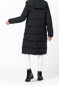 Svart puffjacka med huva, quiltat mönster, långa ärmar och medellång skärning, tillsammans med vita byxor och svarta ankelstövlar.