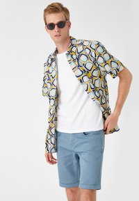 Camisa de botones de manga corta con un patrón geométrico en azul, amarillo y crema, combinada con pantalones cortos azul claro enrollados y gafas de sol oscuras.