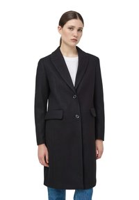 Cappotto di lana nero con maniche lunghe, revers a punta, due tasche frontali e due bottoni. Indossato sopra una maglietta bianca e jeans blu. Texture liscia.