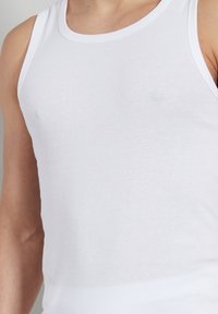 Weißes Tanktop aus geripptem Baumwollstoff, mit breiten Trägern und enger Passform. Der Stoff hat eine glatte Textur ohne Muster oder Verzierungen.
