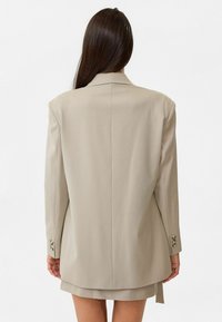 Beige oversized blazer met scherpe schouders, gestructureerde stof, een enkele vent aan de achterkant en knoopmanchetten. Tailored fit met minimale details.
