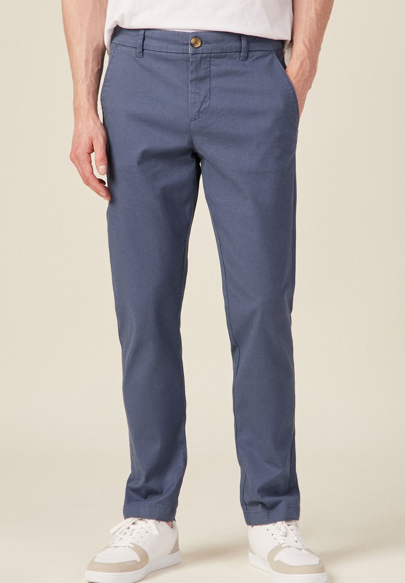 Pantalons droit bleu marine avec une finition texturée, fermeture à bouton et poches latérales ; associés à des baskets blanches avec des accents beiges.