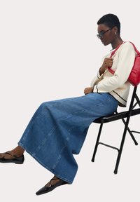 Denim maxikjol med en utsvängd silhuett, ljusblå färg, i kombination med en krämfärgad tröja med svarta detaljer. Svarta flats och en röd väska.