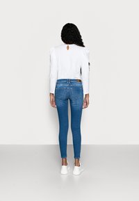 Mujer con cabello largo y rizado, vistiendo una camiseta blanca de manga larga, jeans ajustados azules y zapatillas blancas, de pie frente a una pared en blanco.
