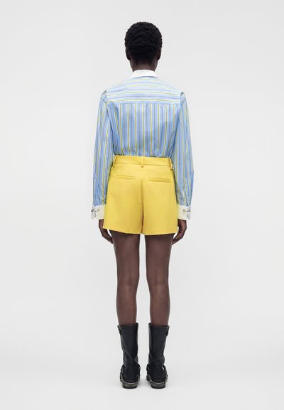 Personne debout de dos, portant un t-shirt à manches longues rayé bleu et jaune, un short jaune et des bottes noires mi-mollet.