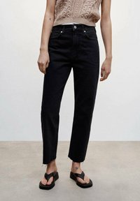 Jeans noirs avec un design à jambe droite, ourlets effilochés et style classique à cinq poches. Associés à un haut en tricot beige et des tongs noires.