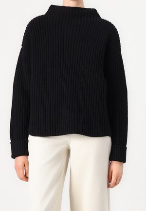 Osoba ubrana w czarny prążkowany sweter z długim rękawem i białe spodnie, stojąca na jednolitym tle.