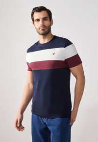 Navy, white, and burgundy striped t-shirt made of cotton. Short sleeves, round neck, with a small bird logo on the chest. Casual fit.