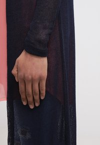 Stickad cardigan i marinblått, med långa ärmar, ett strukturerat mönster och en sidoklyvning. Ett rosa plagg syns under.