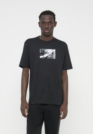 Μαύρο βαμβακερό μπλουζάκι με γραφικό τύπωμα παπουτσιού σε ασπρόμαυρο, πάνω σε υφασμένο φόντο. Κοντά μανίκια, κανονική εφαρμογή.