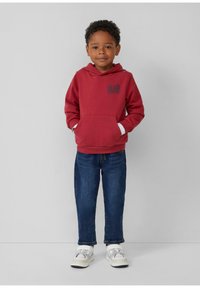 Rode hoodie met een voorzak en bedrukte ontwerp, gecombineerd met blauwe spijkerbroeken en witte sneakers met grijze accenten. Neutrale achtergrond.