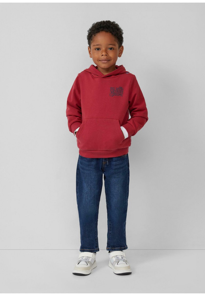 Rode hoodie met een voorzak en bedrukte ontwerp, gecombineerd met blauwe spijkerbroeken en witte sneakers met grijze accenten. Neutrale achtergrond.