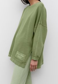 Felpa verde oversize con collo a giro, maniche lunghe e una patch con la scritta "Marc O'Polo PEACE" sul davanti. Realizzata in tessuto morbido.