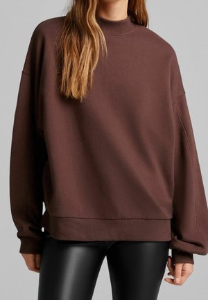 Femme portant un sweat-shirt ample à manches longues marron associé à un pantalon en cuir noir, vue du cou aux hanches sur fond gris.