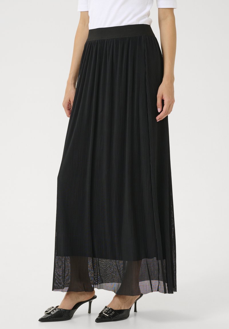 Jupe maxi plissée noire avec une taille ajustée, superposition transparente et tissu texturé. Portée avec des mules à talons décoratives noires.