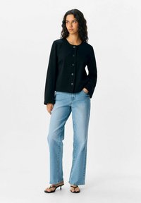 Giacca nera corta con scollo rotondo e sei bottoni neri, abbinata a jeans dritti azzurri e sandali neri con tacco.