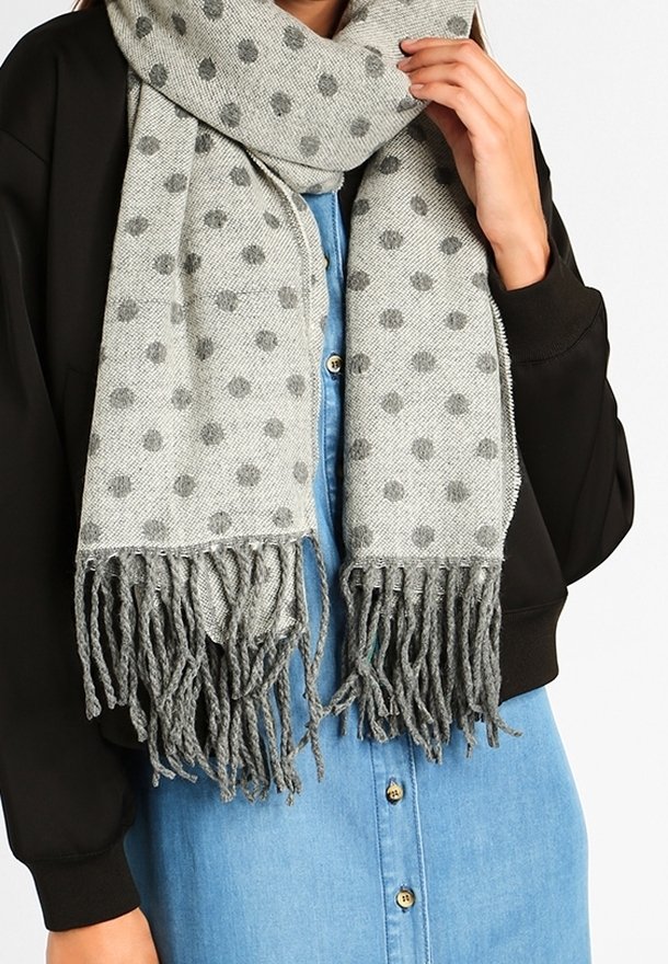 Écharpe grise à pois foncés avec des extrémités à franges, portée sur une veste noire et une robe bleue boutonnée.