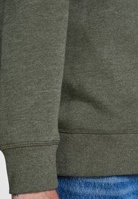 Detalle de sudadera verde oliva con tejido texturizado, puños y bajo acanalados. Pantalones vaqueros azules visibles en la parte inferior.