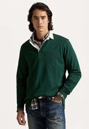Donkergroene longsleeve polo met een witte kraag, voorzien van een klein logo. Gelaagd met een geruit overhemd en gedragen met een blauwe spijkerbroek.