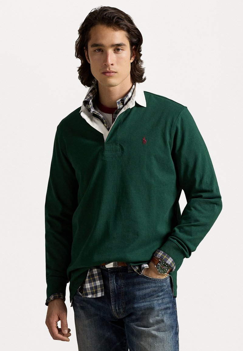 Donkergroene longsleeve polo met een witte kraag, voorzien van een klein logo. Gelaagd met een geruit overhemd en gedragen met een blauwe spijkerbroek.